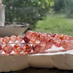 Puede incluir: Dos pulseras de cuentas redondas translúcidas con vetas rojas y blancas. Las cuentas descansan sobre una superficie blanca y texturizada. El fondo incluye una vista borrosa de una ventana y un cuenco de cerámica blanco.