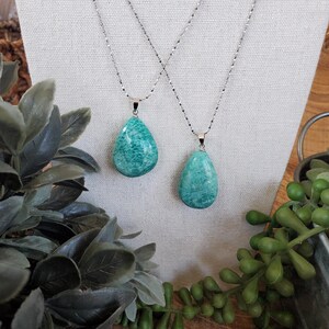 May include: Two Amazonite teardrop pendant necklaces with silver chains. The pendants are a vibrant teal colour with natural marbling. The necklaces are displayed on a cream-coloured fabric, with greenery in the foreground and background.