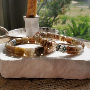 May include: Two bracelets made of rectangular, translucent beads in shades of brown, tan, and gray. The bracelets are resting on a white, textured surface. A candle and a decorative dish are in the background.