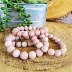 Puede incluir: Tres pulseras de piedras preciosas rosas hechas de cuentas redondas. Las pulseras est&aacute;n apiladas una encima de la otra sobre una superficie de madera.