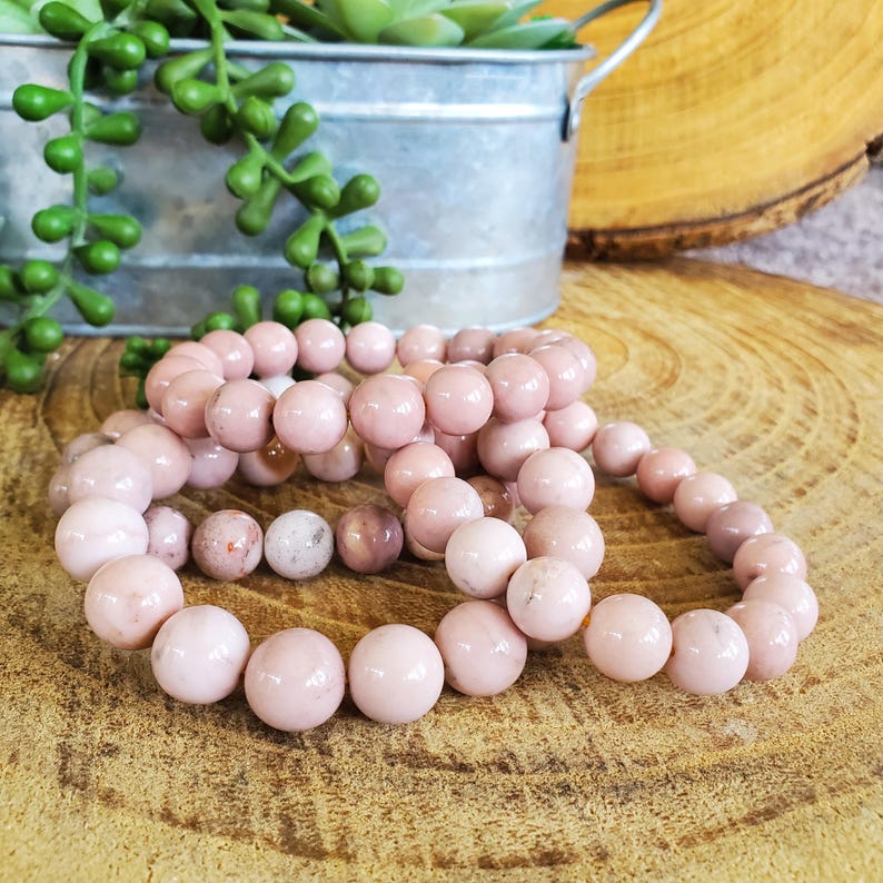 Puede incluir: Tres pulseras de piedras preciosas rosas hechas de cuentas redondas. Las pulseras est&aacute;n apiladas una encima de la otra sobre una superficie de madera.