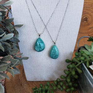 May include: Two necklaces with teal teardrop pendants suspended from delicate silver chains. The pendants have a mottled appearance. The necklaces are displayed on a white fabric stand, with greenery in the background.
