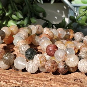 May include: A close-up of a strand of gemstone beads. The beads are a mix of clear, tan, brown, and black colors. The beads are round and appear to be made of a natural stone material. The beads are strung together on a string.