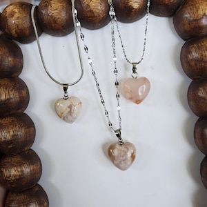 Op de afbeelding: Drie kettingen met hartvormige hangers. De hangers zijn gemaakt van steen in beige, roze en witte tinten. De kettingen zijn zilverkleurig en hebben verschillende ontwerpen. De kettingen worden gepresenteerd tegen een witte achtergrond.