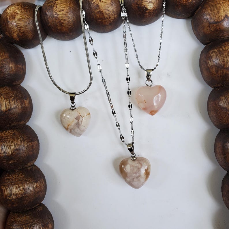 Op de afbeelding: Drie kettingen met hartvormige hangers. De hangers zijn gemaakt van steen in beige, roze en witte tinten. De kettingen zijn zilverkleurig en hebben verschillende ontwerpen. De kettingen worden gepresenteerd tegen een witte achtergrond.