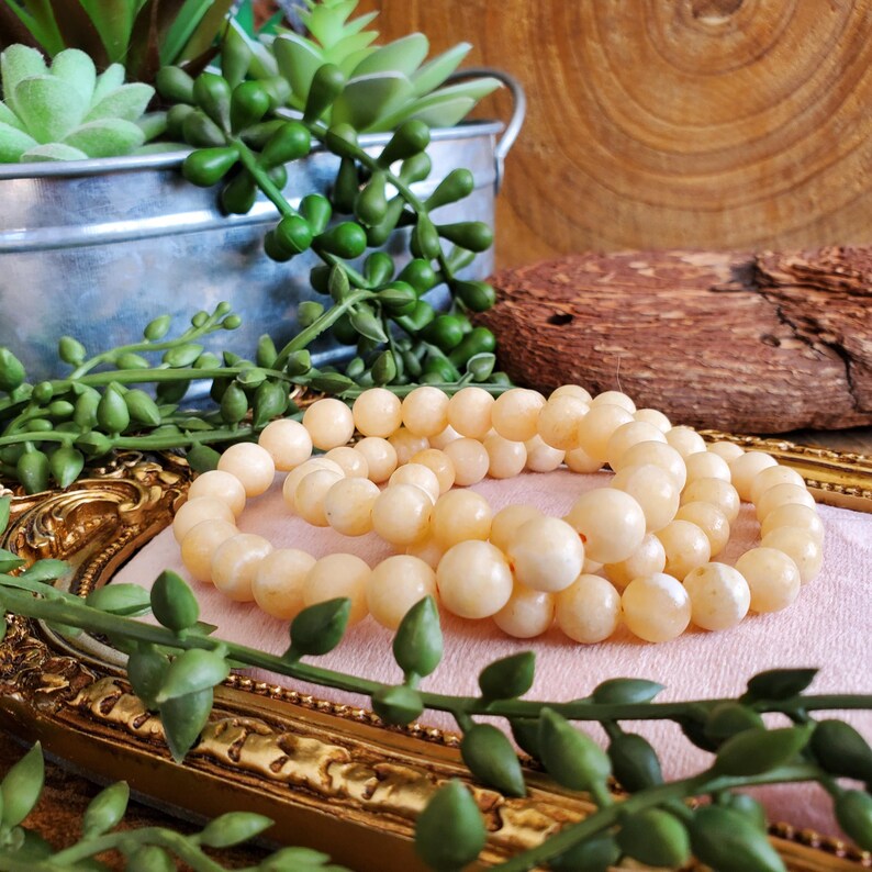 May include: Three strands of pale yellow beaded bracelets are displayed on a pink surface within a gold-framed setting. The bracelets are surrounded by green leafy plants and a small metal bucket with greenery.