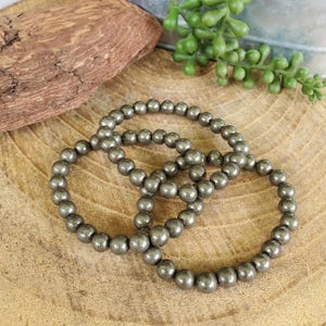 May include: Three beaded bracelets made of small, round, metallic gray beads. The bracelets are arranged in a circular pattern on a wooden surface. The beads have a slightly textured appearance, and the overall aesthetic is earthy and natural.