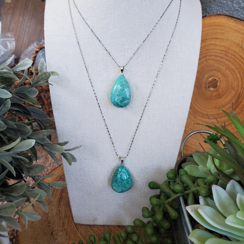 May include: Two silver necklaces, each with a turquoise teardrop pendant. The pendants are displayed on a white fabric surface, with greenery and a wooden background. The necklaces are of different lengths.