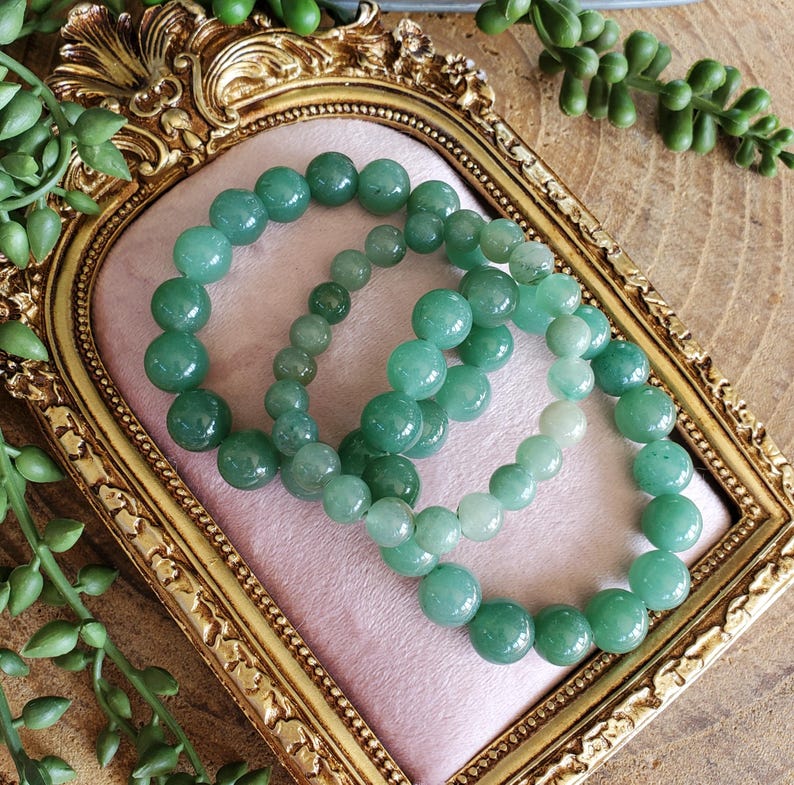 May include: Three green beaded bracelets of varying sizes rest on a pink surface within a gold-framed mirror. The beads are a consistent shade of green, and the bracelets are arranged to showcase their different sizes. The background includes greenery.