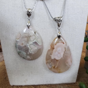 May include: Two teardrop-shaped pendants with silver chains. The pendants are made of polished stone, featuring a mix of pink, white, and gray patterns. Each pendant is suspended from a decorative silver bail.