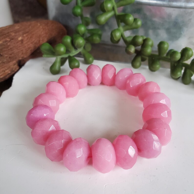 May include: A pink beaded bracelet with faceted, oval-shaped beads. The bracelet is a light pink color and is resting on a white surface. Green plant stems are in the background.