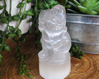 May include: A translucent, carved figurine of a seated figure with a beard and hat, possibly a gnome or Santa Claus. The figurine is mounted on a cylindrical base. The sculpture is made of a light-coloured, possibly crystal, material.