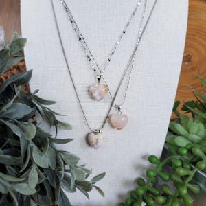 Op de afbeelding: Drie kettingen met hartvormige hangers. De hangers zijn een mix van roze en cr&egrave;mekleur. De kettingen zijn zilverkleurig en vari&euml;ren in stijl. De kettingen worden gepresenteerd op een witte stoffen ondergrond, met groen op de voor- en achtergrond.
