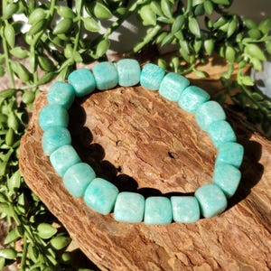 Puede incluir: Un brazalete de Amazonita turquesa. El brazalete est&aacute; hecho de cuentas cuadradas, cada una con un patr&oacute;n &uacute;nico. Las cuentas est&aacute;n ensartadas, formando una forma circular. El brazalete se exhibe sobre un trozo de madera, con follaje verde en el fondo.