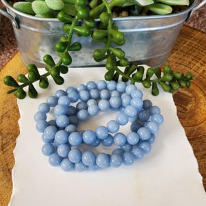 May include: A strand of light blue round beads, likely gemstones, resting on a white surface. The beads are a consistent size and color, with a smooth, polished appearance. The background includes a metal container with green plants and a wooden surface.