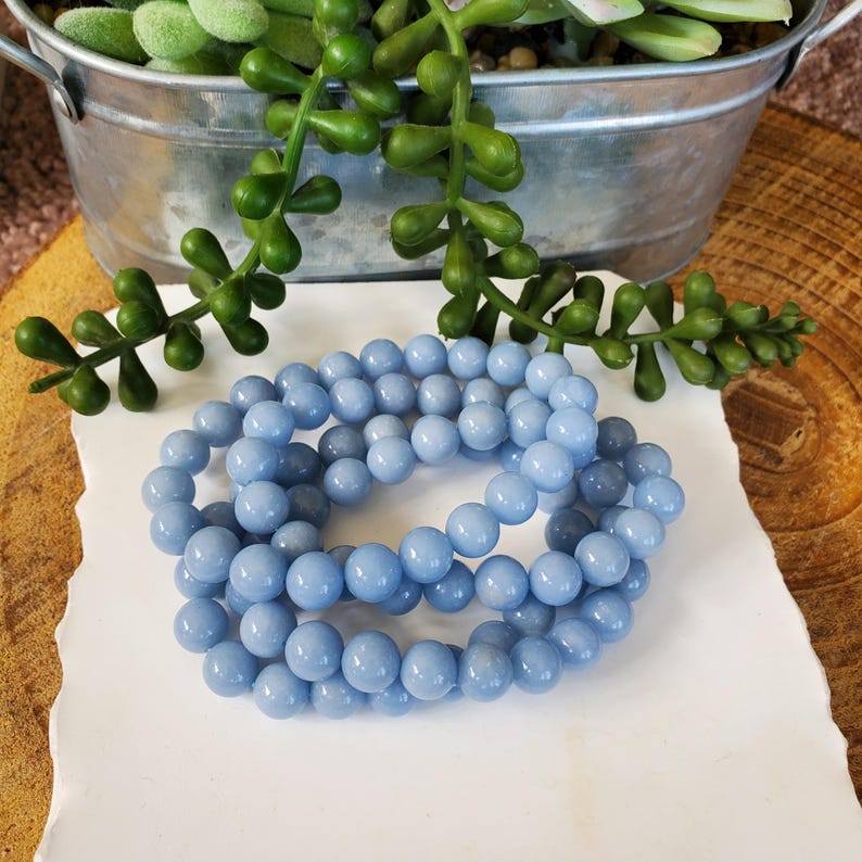 May include: A strand of light blue round beads, likely gemstones, resting on a white surface. The beads are a consistent size and color, with a smooth, polished appearance. The background includes a metal container with green plants and a wooden surface.
