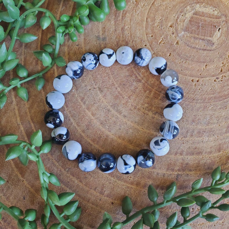 May include: A beaded bracelet featuring round, white and black patterned beads. The bracelet is displayed on a wooden surface, surrounded by green leafy sprigs.