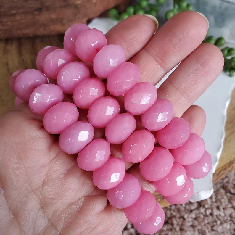 May include: A strand of faceted, oval-shaped pink beads. The beads have a polished, glossy finish and are strung together. The pink color is a soft, pastel hue. The beads are held in a hand, suggesting they are ready to be used for jewelry making.
