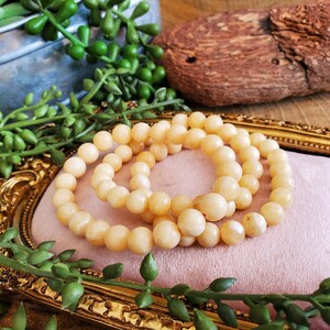 May include: Three strands of light peach-colored beaded necklaces are arranged on a pink surface within a gold-framed tray. The beads are round and uniform in size. Green leafy plants and a piece of wood are in the background.