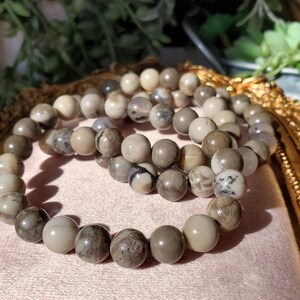 May include: Three beaded bracelets with natural stone beads in shades of gray, brown, and cream. The round beads have unique patterns and textures. The bracelets are displayed on a pink surface.