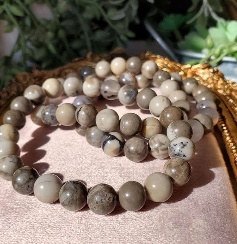 May include: Three beaded bracelets with natural stone beads in shades of gray, brown, and cream. The round beads have unique patterns and textures. The bracelets are displayed on a pink surface.