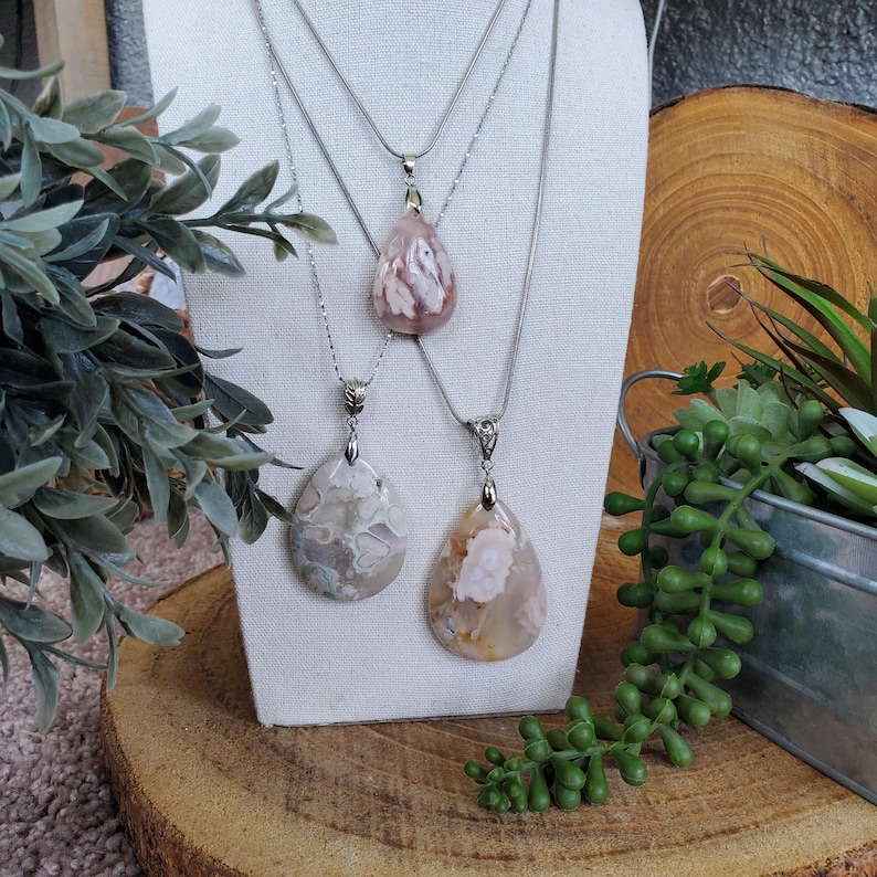 May include: Three silver necklaces with teardrop-shaped pendants. The pendants are made of polished stone in shades of pink, cream, and gray. The necklaces are displayed on a white stand, with greenery and a wooden slice in the background.