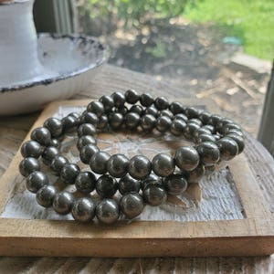 May include: Two bracelets made of dark gray, spherical beads. The bracelets are resting on a light brown, wooden tray with a white border. The beads have a metallic sheen, and the bracelets are stacked on top of each other.