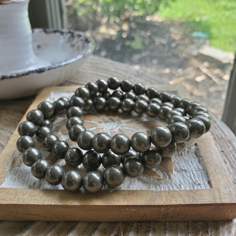 May include: Two bracelets made of dark gray, spherical beads. The bracelets are resting on a light brown, wooden tray with a white border. The beads have a metallic sheen, and the bracelets are stacked on top of each other.