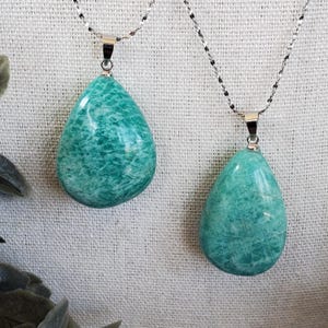 May include: Two turquoise-green teardrop pendants on silver chains. The pendants have a marbled effect. The background is a textured, off-white fabric. Each pendant is a unique piece of jewellery.