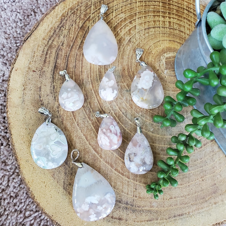May include: A collection of eight teardrop-shaped pendants with silver-colored bails. The pendants are made of pink and white patterned stone. The pendants are displayed on a wooden surface with a small metal bucket and greenery in the background.