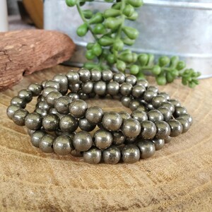 May include: Three strands of pyrite gemstone beads, each with a metallic, golden-gray luster. The beads are round and appear to be strung together, creating a bracelet or necklace. The jewelry is displayed on a wooden surface.