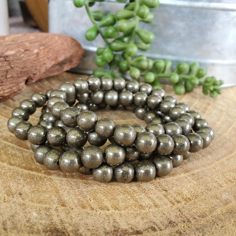 May include: Three strands of pyrite gemstone beads, each with a metallic, golden-gray luster. The beads are round and appear to be strung together, creating a bracelet or necklace. The jewelry is displayed on a wooden surface.
