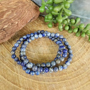 May include: Three blue beaded bracelets with a mix of dark and light blue beads. The bracelets are arranged on a wooden surface, with a piece of wood and green plant in the background.