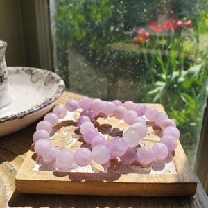 K&ouml;nnte beinhalten: Zwei hellviolette Perlenarmb&auml;nder liegen auf einem kleinen, hellbraunen Holztablett. Die Armb&auml;nder bestehen aus runden, polierten Perlen. Der Hintergrund zeigt ein Fenster mit Blick auf Gr&uuml;n und Blumen.
