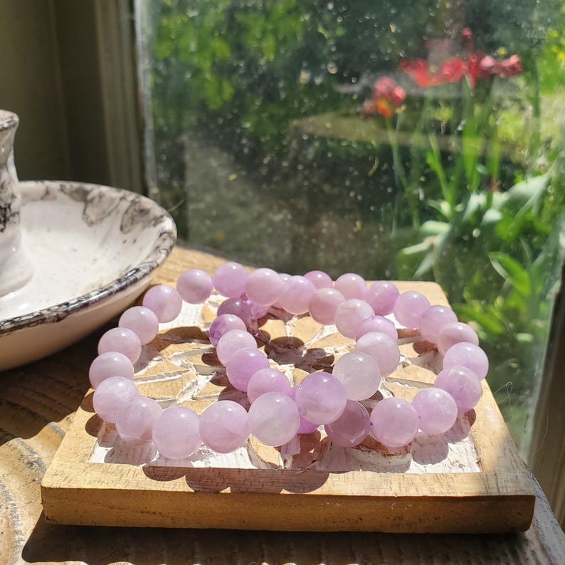 K&ouml;nnte beinhalten: Zwei hellviolette Perlenarmb&auml;nder liegen auf einem kleinen, hellbraunen Holztablett. Die Armb&auml;nder bestehen aus runden, polierten Perlen. Der Hintergrund zeigt ein Fenster mit Blick auf Gr&uuml;n und Blumen.