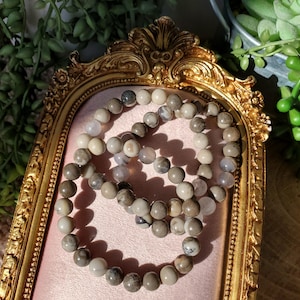 May include: Beaded bracelets with a mix of gray, white, and tan stones are displayed on a gold-framed, ornate mirror. The bracelets are arranged on a pink surface, with greenery in the background. The beads appear to be approximately 0.3 inches in diameter.