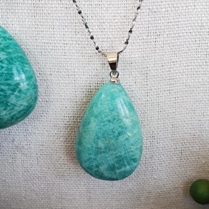 May include: A turquoise teardrop pendant necklace with a silver chain. The pendant is a polished stone with a smooth surface. The necklace is displayed against a white textured background.