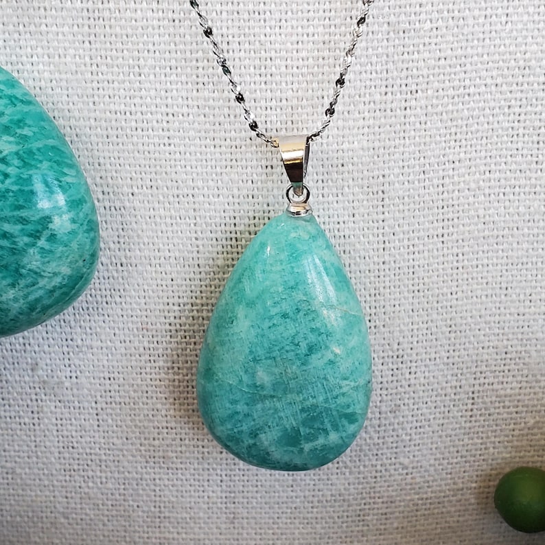 May include: A turquoise teardrop pendant necklace with a silver chain. The pendant is a polished stone with a smooth surface. The necklace is displayed against a white textured background.