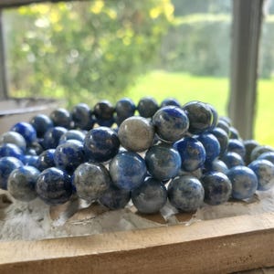 May include: A stack of round, blue and gray gemstone bead bracelets. The beads have a marbled appearance, with varying shades of blue and gray. The bracelets are displayed on a light-colored, textured surface, with a blurred green background.