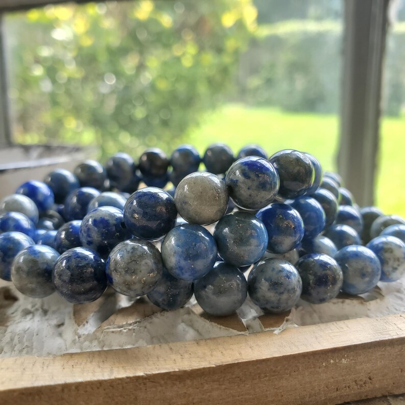 May include: A stack of round, blue and gray gemstone bead bracelets. The beads have a marbled appearance, with varying shades of blue and gray. The bracelets are displayed on a light-colored, textured surface, with a blurred green background.