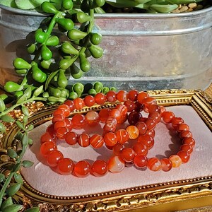 May include: Three bracelets made of polished, round, orange-red carnelian beads. The beads have white and orange banding. The bracelets are displayed on a pink surface inside a gold-coloured frame, with greenery in the background.