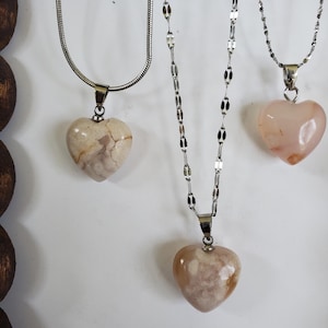 Op de afbeelding: Drie kettingen met hartvormige hangers. De hangers zijn gemaakt van steen in beige en roze tinten. De kettingen zijn zilverkleurig en vari&euml;ren in stijl. De kettingen worden gepresenteerd tegen een witte achtergrond.