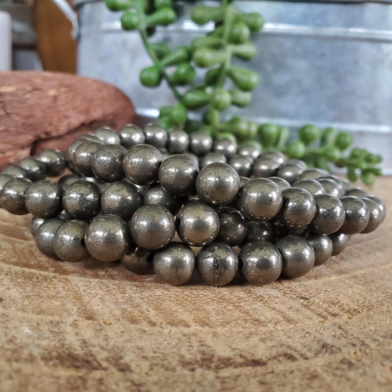 May include: A stack of three bracelets made of metallic gray pyrite beads. The beads are round and have a slightly textured surface. The bracelets are displayed on a wooden surface, with a metal container and greenery in the background.