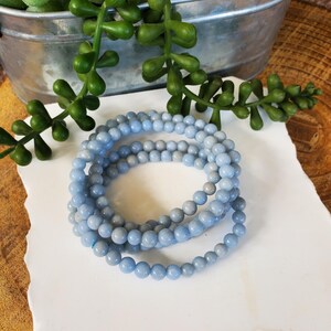 May include: Three strands of light blue beaded bracelets are coiled on a white surface. The beads are small and round. Green leafy plants and a metal container are in the background.