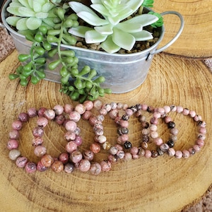 Puede incluir: Cuatro pulseras de piedras preciosas rosadas y blancas con cuentas redondas. Las pulseras est&aacute;n apiladas una encima de la otra sobre una superficie de madera.