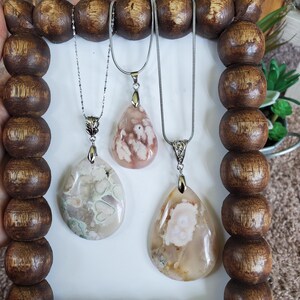 May include: Three gemstone pendant necklaces with silver chains. The pendants are teardrop and oval shaped, featuring shades of cream, pink, and brown. The necklaces are displayed on a wooden beaded frame.