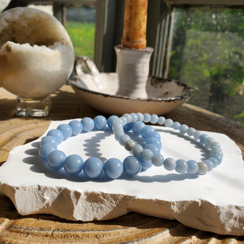 May include: Two light blue beaded bracelets rest on a white, textured surface. The bracelets are made of round, polished beads. A candle in a white holder and a geode sphere are in the background.