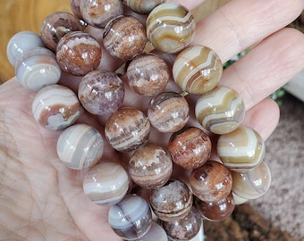 Puede incluir: Una pulsera compuesta por múltiples cuentas esféricas pulidas de ágata bandeada. Las cuentas presentan una variedad de tonos tierra, incluyendo matices de marrón, bronceado, crema y blanco, con patrones circulares concéntricos visibles.