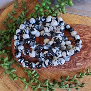 May include: Several beaded bracelets with black and white patterned beads are arranged on a wooden surface. The bracelets are made of round, polished beads. Greenery surrounds the bracelets, adding a natural element to the composition.