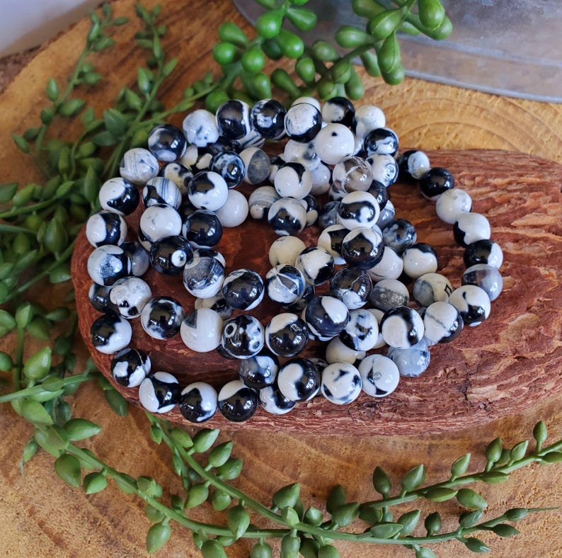 May include: Several beaded bracelets with black and white patterned beads are arranged on a wooden surface. The bracelets are made of round, polished beads. Greenery surrounds the bracelets, adding a natural element to the composition.